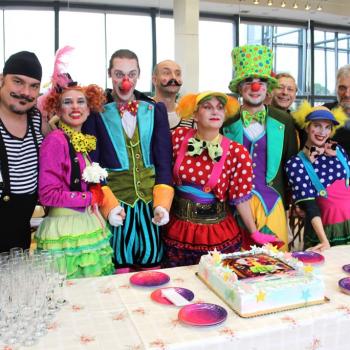 Aktorzy w kolorowych strojach stoją za stołem na którym znajduje się tort, kieliszki i butelki szampana.