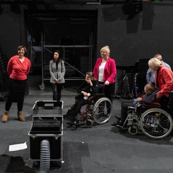 Dwie osoby na wózkach inwalidzkich wraz z opiekunami i pracownikami znajdują się na scenie