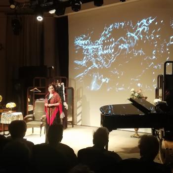 Magdalena Tomaszewska stoi na scenie. Po prawej stronie czarny fortepian. W tle plansza z wizualizacją w kształcie fal.