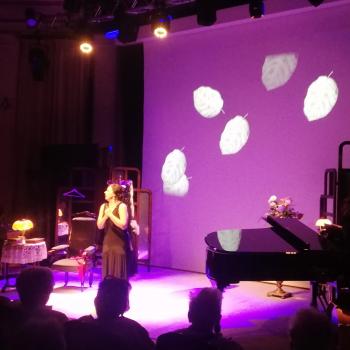 Magdalena Tomaszewska w czarnej sukni stoi na scenie. Po prawej stronie czarny fortepian. W tle plansza z wizualizacją w kształcie opadających liści.