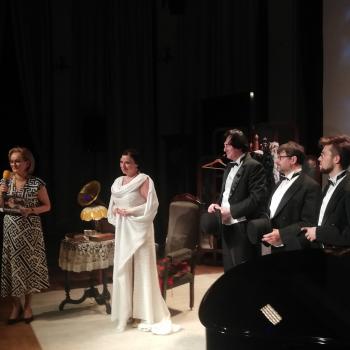 Magdalena Tomaszewska w białej sukni stoi na scenie, po prawej trzech mężczyzn w smokingach, po lewej kobieta trzymająca w dłoni mikrofon.