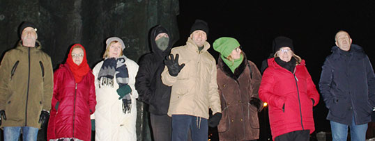 Scena zbiorowa. Osiem osób patrzących w kamerę, w tle fragment pogrążonego w półmroku pomnika Broniewskiego