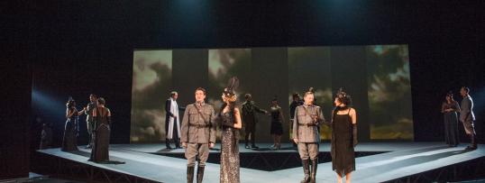 Mężczyźni w mundurach, kobiety w sukniach wieczorowych stojący na scenie. Fotografia z przedstawienia Gwiazdy rdzewieją na dnie Wisły.