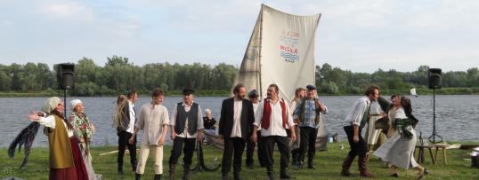 Aktorzy stoją na brzegu Wisły porośniętym trawą. W tle maszt i żagiel łodzi z nadrukiem Wisła 2017