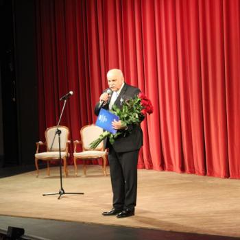 Duża Scena płockiego teatru. Na czerwonej kurtynie Krzysztof Szuster z kwiatami przemawiający do mikrofonu.