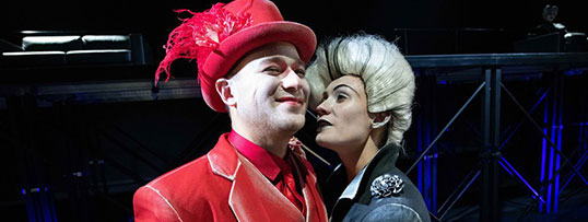 Aleksander Maciejczyk (Lekkoduch) w czerwonym garniturze  i kapeluszu, obok Maja Rybicka (Rena) w czarnej sukni i siwej peruce, zwróceni blisko do siebie twarzami.