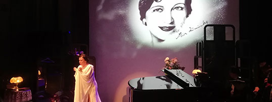 Magdalena Tomaszewska w białej sukni stoi na scenie. Po prawej stronie czarny fortepian. W tle plansza z wizualizacją twarzy Miry Zimińskiej