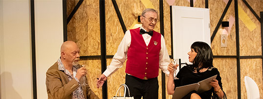 Centralną postacią jest Jerzy Bończak ubrany w czerwoną, hotelową kamizelkę. Stoi. Po lewej stronie siedzi Marek Walczak, po prawej Magdalena Tomaszewska
