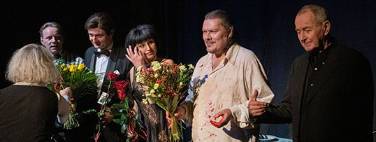 Aktorzy stoją na scenie i trzymają w dłoniach kwiaty. Po prawej dyrektor Marek Mokrowiecki Czarne tło.