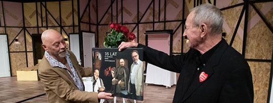 Dwaj mężczyźni stoją na scenie. Po lewej Marek Walczak skierowany w prawą stronę trzymający w prawej dłoni pamiątkowy obraz odbierany przez dyrektora Marka Mokrowieckiego stojącego z prawej strony w czarnym garniturze. Zza obrazu widać trzymany przez Marka w drugiej dłoni bukiet czerwonych róż.