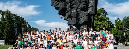 Zdjęcie grupowe. Grupa kilkudziesięciu osób stoi na schodach prowadzących pod pomnik Broniewskiego. W tle pomnik na tle błękitnego nieba.